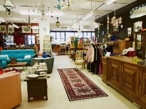 Eine Holzkommode, Spiegel, ein Teppich und eine Kleiderstange mit Kleidung auf den Bügeln, ein Sofa und Lampen hängen und stehen in einem Geschäft