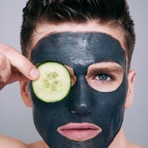 Ein Mann hat eine dunkle Gesichtsmaske im Gesicht und hält sich eine Gurkenscheibe an das rechte Auge