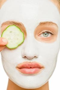 Eine Frau hat eine weiße Gesichtsmaske im Gesicht und hält sich eine Gurkenscheibe an das rechte Auge