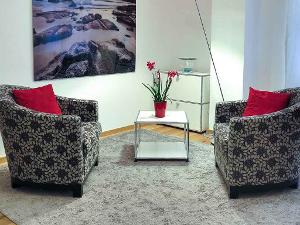 Besprechungszimmer mit zwei grauen Sesseln, einem weißen Tisch mit roten Blumen in einer roten Blumenvase und einem Bild an der Wand.