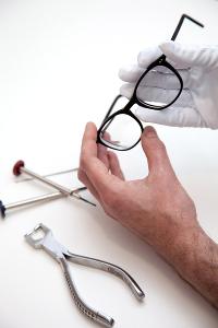Ein Mensch hat weiße Schutzhandschuhe an und schaut sich eine Brille an, auf dem Tisch liegt Werkzeug für die Reparatur
