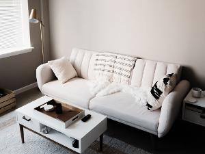 Ein weißes Sofa steht an einer Wand, davor ein weißer Wohnzimmertisch und auf dem Sofa liegen eine Decke und zwei Kissen