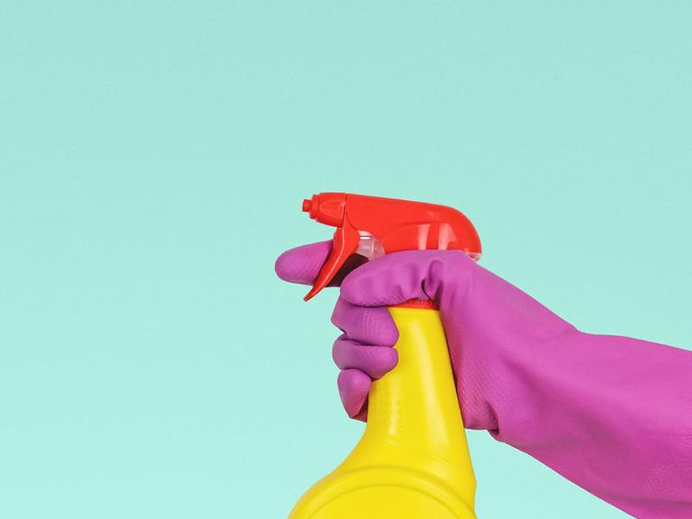 Ein lilafarbener Handschuh hält eine gelbe Sprühflasche mit rotem Verschluss in der Hand, vor einem mintgrünen Hintergrund