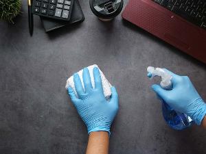 Zwei blaue Handschuhe, in der einen Hand einen Schwamm, in der anderen Hand eine Sprühflasche und bearbeitet eine Schreibtischunterlage