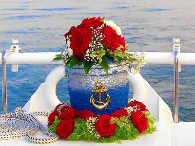 Ein blau-silbernes Gefäß umringt von einem Blumenkranz mit roten Blumen, auf dem Gefäß liegen auch rote und weiße Blumen- alles steht vorne an einem weißem Schiff und man sieht noch das Wasser