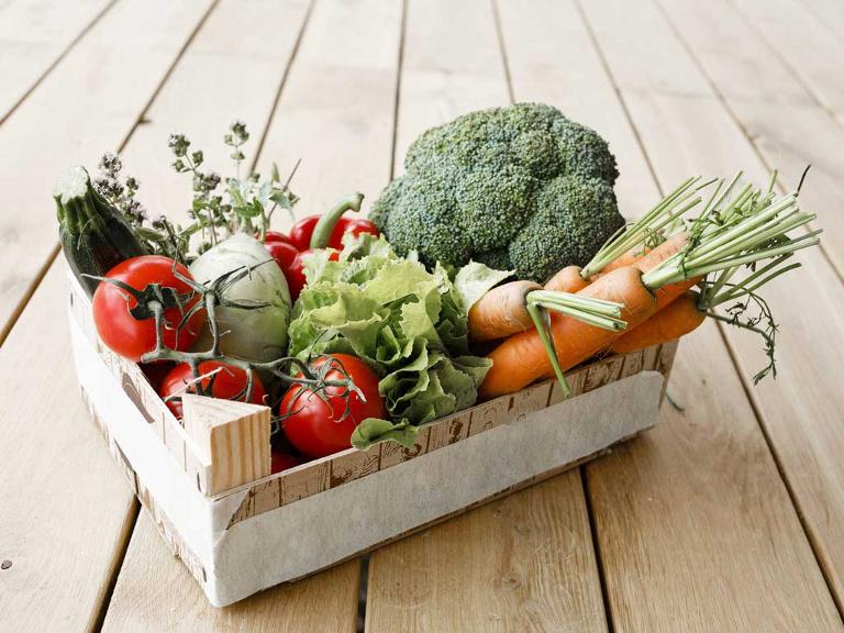 Eine Holzkiste auf einem Holzfußboden gefüllt mit Broccoli, Karotten, Tomaten, Salat, Paprika und Kohlrabi