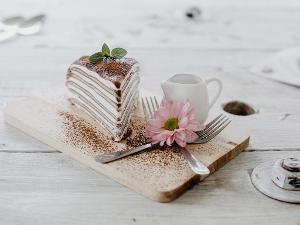 ein Stück Kuchen mit Kakaopulver bestreut, auf einem holzbrettschen, zwei gabeln halten eine rosafarbene Blüte und ein Milchkännchen steht an der Seite