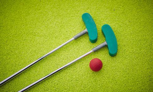 Zwei grüne Minigolfschläger mit einem roten Golfball auf einem Rasen liegend