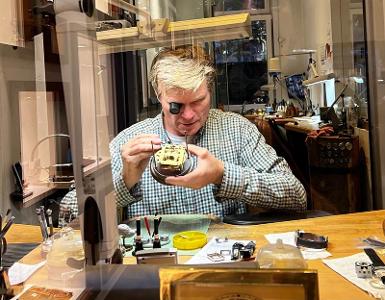 Ein Mann mit Lupe sitzt an einem Tisch und repariert eine Uhr