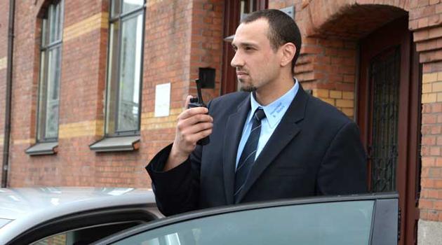 Mann im dunklen Anzug steht vor offener Autotür mit Funkgerät in der Hand.