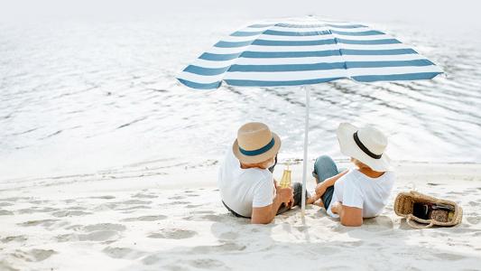 Zwei Menschen haben einen Hut auf und liegen im Sand unter einem Sonnenschirm