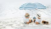 Zwei Menschen haben einen Hut auf und liegen im Sand unter einem Sonnenschirm