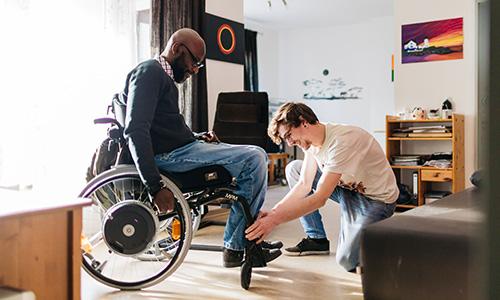 Ein Mann hilft einem anderen, der in einem Rollstuhl sitz- die Füße abzustellen