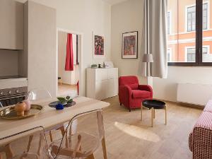 Ein heller Raum mit Küchenzeile, Backofen, einem Esstisch mit Stühlen aus Holz und einem roten Sessel mit Beistelltisch vor einem Fenster