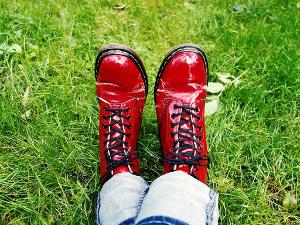 Übereinandergeschlagene Beine mit einer Jeans liegen auf einer grünen Wiese und rote Stiefel mit schwarzer Sohle und schwarzem Band an den Füßen