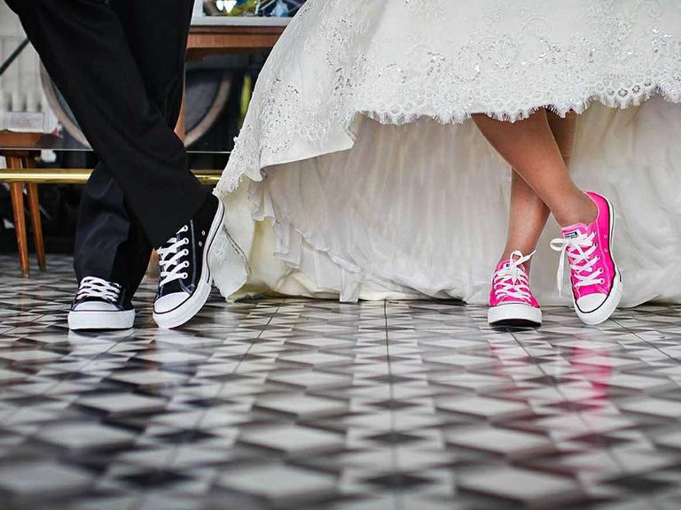 Ein Mann trägt schwarze Freizeitschuhe mit weißem Band, eine Frau trägt die gleichen Schuhe nur in pink, bei beiden sieht man nur den unteren Teil des Körpers- sie trägt ein weißes Brautkleid, er einen Anzug in schwarz