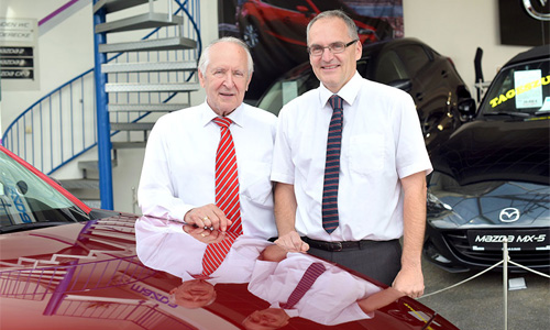 Zwei Männer in weißen Hemden und Krawatte stehen hinter einem roten Auto und lächeln in die Kamera
