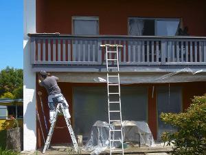 Ein Mensch sitzt auf einer Leiter während er die Balkonbrüstung eines zweistöckigen Hauses anmalt