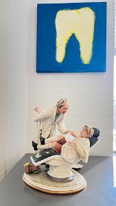 Eine Dekofigur mit einem Menschen auf einem Zahnarztstuhl und einem anderen der bereit ist den Patienten zu untersuchen, im Hintergrund hängt ein Bild mit einem Zahn an der weißen Wand