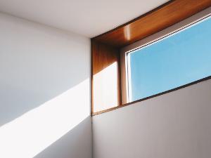 Ein Glasfenster mit braunem Rahmen eingebaut in eine weiße Wand, man sieht einen blauen Himmel und die Sonne wirft einen Lichtstrahl an die Wand