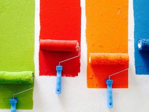 Vier Farbrollen liegen nebeneinander, eine grüne, eine rote, eine orange und eine blaue- jede malt einen Strich an die weiße Wand