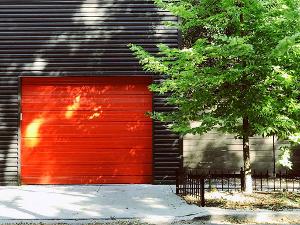 Ein rotes Garagentor in einer dunklen Häuserwand und rechts daneben eine Baum mit grünen Blättern