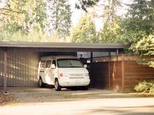 Ein Carport aus Holz vor einem Wald und darunter steht ein weißes Auto