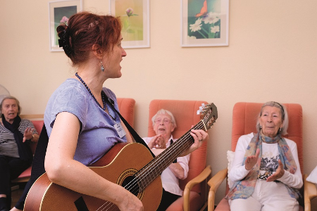 Senioren sitzen auf Stühlen und klatschen in die Hände- eine Frau mit Gitarre steht vor ihnen und singt