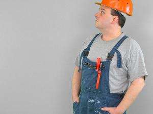 Ein Mann mit grauem T-Shirt und darüber einen Blaumann mit Werkzeug in der Vordertasche schaut nach oben und hat einen orangefarbenen Helm auf und die Hände in den Hosentaschen