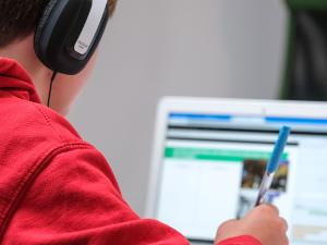 Ein Mensch in einem roten Pullover und mit einem Kopfhörer auf den Ohren, sitzt vor einem Laptop