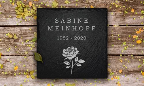 Gedenktafeln aus Schiefer mit weißer Schrift und einer Rose, liegt auf Holzpaneelen mit Blüten darauf liegend