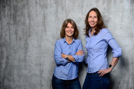 Zwei Frauen in Hemd und Blazer vor grauem Hintergrund