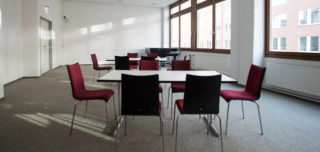 Ein kleiner Raum mit zwei weißem Tischen an denen jeweils sechs rote Stühle stehen