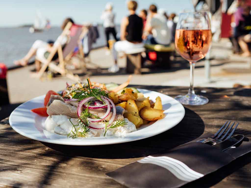 Teller mit Fisch und Bratkartoffeln und einem Glas Wein und im Hintergrund die Elbe mit vielen Gästen.