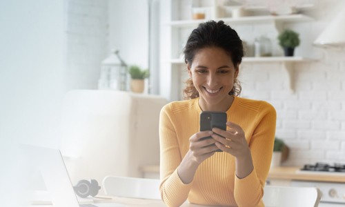 Eine Frau sitzt an einem Tisch, lächelt und schaut auf das Handy in ihren Händen