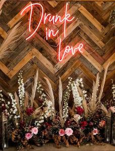 Eine Holzwand mit pinker Leuchtschrift Drunk in Love darauf und davor Blumen