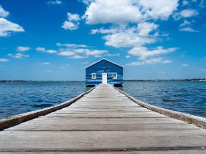 Ein grauer Holzsteg führt auf ein blaues Haus mit weißer Tür auf dem Wasser