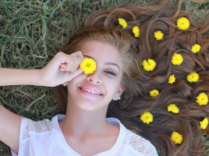 Ein Mädchen liegt auf einer Wiese, auf ihren Haaren liegen gelbe Blüten, sie hält sich eine gelbe Blüte vor ihr Auge und lächelt in die Kamera