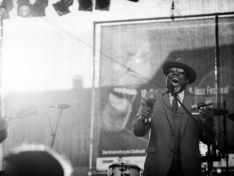 Ein schwarz-weiss Foto auf dem ein Mann mit Sonnenbrille und Hut auf einer Bühne steht und singt