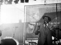 Ein schwarz-weiss Foto auf dem ein Mann mit Sonnenbrille und Hut auf einer Bühne steht und singt