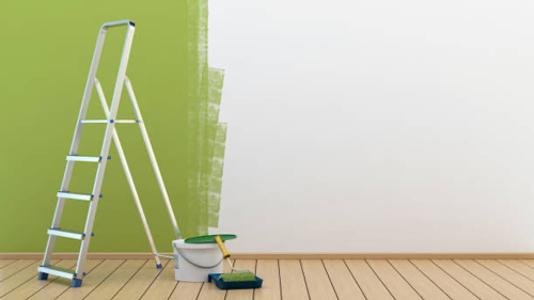 Eine weiße Wand zur Hälfte grün davor steht eine Leiter mit einem Farbtopf