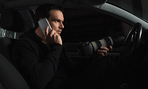 Mann mit Handy am Ohr und Kamera in der Hand im Auto bei einer Ermittlung.