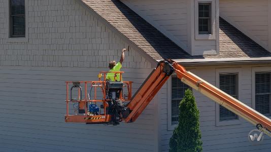 Ein Mensch steht auf der Hebebühne eines Krans und malt das Dach an eines Hauses an
