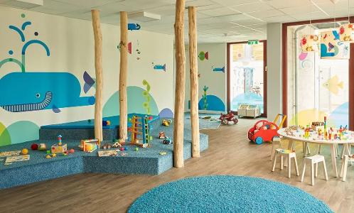 Ein großer heller Raum mit einem blauen Wal an der Wand, Holzstämmen im Raum und eine mit Teppich belegte Anhöhe zum spielen