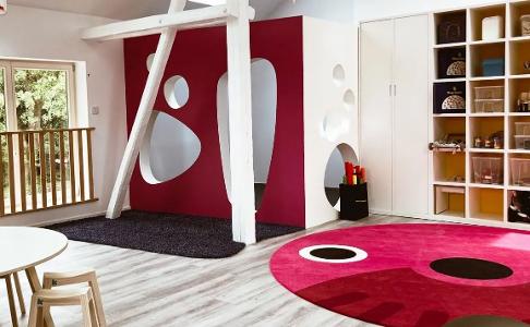 Spielzimmer der Kita kinderzimmer Bergstedter Scheune, mit einem großen, runden, roten Teppich mit Nase und Augen auf einem hellen Holzfußboden, einem Regal aus Holz und Fenstern