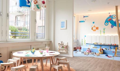 Ein heller Raum in der Kita kinderzimmer Dorotheenstraße mit bunten Tieren an den Wänden, einem Bereich mit blauem Teppich zum spielen und einer kleinen Sitzgruppe aus Holz