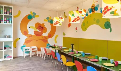 Der Raum zum Essen in der Kita kinderzimmer Klövensteen, bunte Stühle an bunten Tischen, an der Wand ist ein Affe und Bananen gemalt und von der Decke hängen bunte Lampen