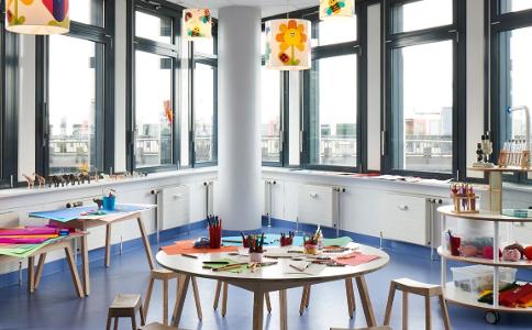 Ein Raum mit Fensterfront, blauem Fußboden und kleinen Tischen mit Stühlen daran in der Kita kinderzimmer Stubbenhuk