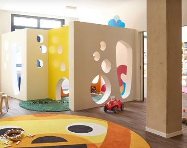Ein Bereich zum spielen in der Kita kinderzimmer Marmelade, ein großer, runder, gelber Teppich liegt auf dem Holzfußboden und Wände mit Löchern teilen den Raum