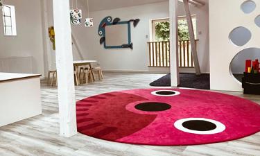 Spielzimmer der Kita kinderzimmer Bergstedter Scheune, mit einem großen, runden, roten Teppich mit Nase und Augen auf einem hellen Holzfußboden